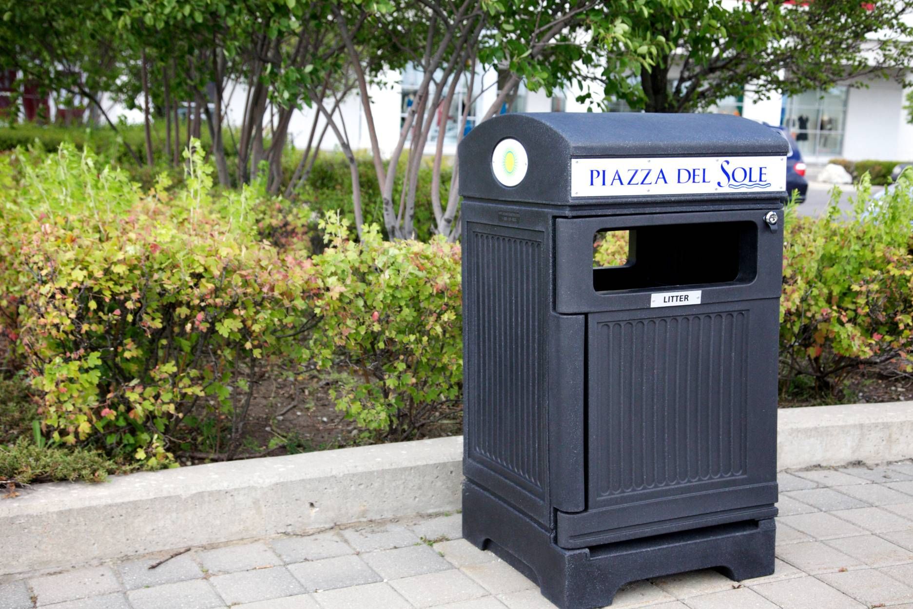 Commercial outdoor waste receptacle in landscaped plaza with durable site furniture for public spaces.